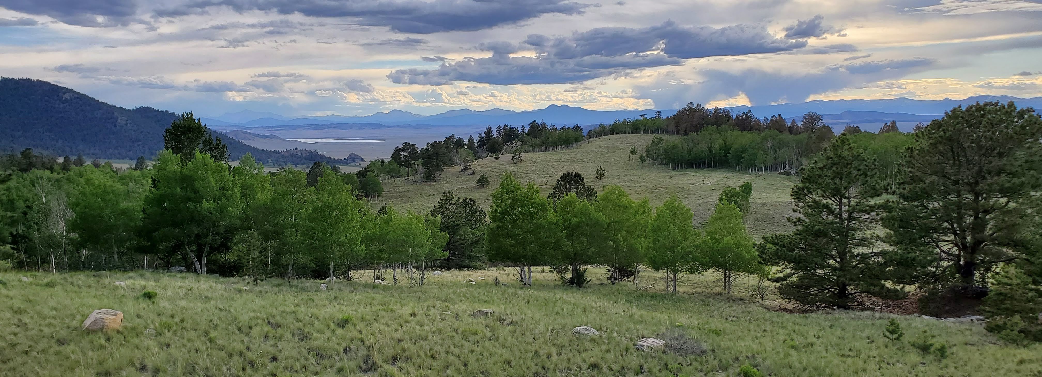 Scenic view of hills and mountains west of Pikes Peak and front range. There are trees and grass rolling across the hills up to distant mountains and the clouds are blue